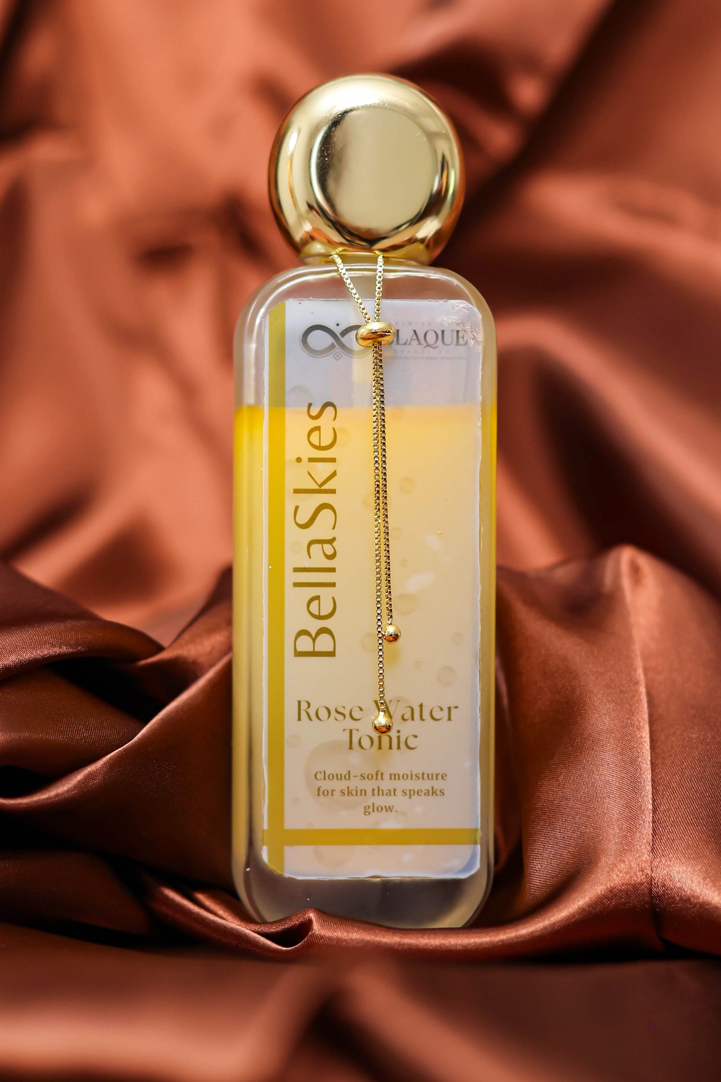 Bella Skies Rose Water Tonic bottle with gold cap on a brown satin background
